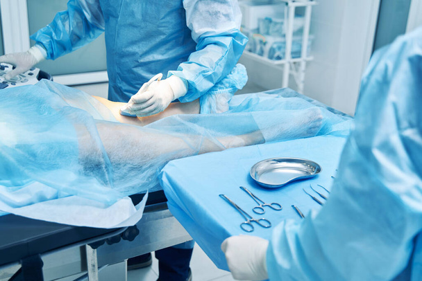 Μη αναγνωρισμένος γιατρός που χρησιμοποιεί συνδεδεμένο μορφοτροπέα στο πόδι ασθενούς ενώ ο συνάδελφός του στέκεται με χειρουργικά εργαλεία - Φωτογραφία, εικόνα