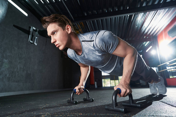 Strong muscular young male athlete performing push-ups on handles during gym workout - Foto, Bild