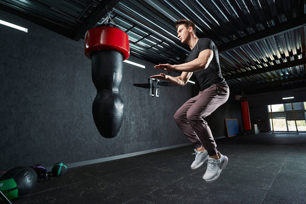 Professional athlete performing tuck jumps in front of hanging punching bag at gym - 写真・画像