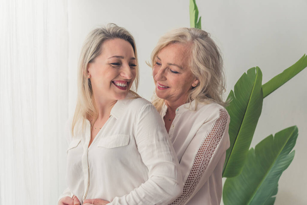 One woman with wavy blonde hair standing in the back hugging another middle-aged caucasian woman. Both laughing and having fun in an elegant apartment. High quality photo - Foto, imagen
