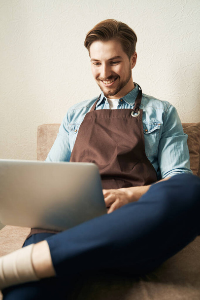 Smiling cheerful male sitting on sofa in room and typing on his laptop - Photo, image