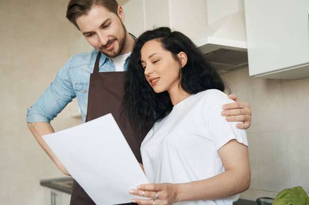 Waist-up portrait of smiling woman and pleased man standing in kitchen and reading documentation - Photo, Image