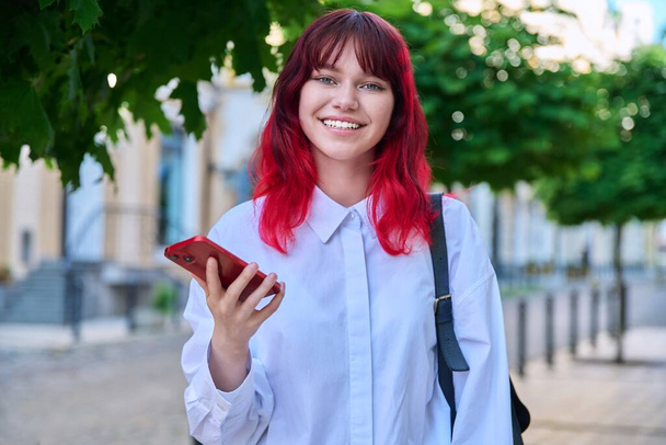 Outdoor portrait of fashionable teenage female student with smartphone in hands looking at camera, on city. Teenager with trendy colored hairstyle red hair. Youth fashion, beauty, technology lifestyle - Фото, зображення