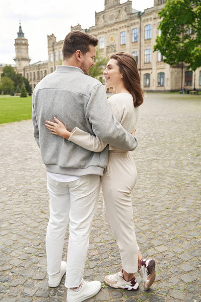 Comprimento total do homem abraçando a mulher e sorrindo enquanto está em pé na avenida ao ar livre - Foto, Imagem