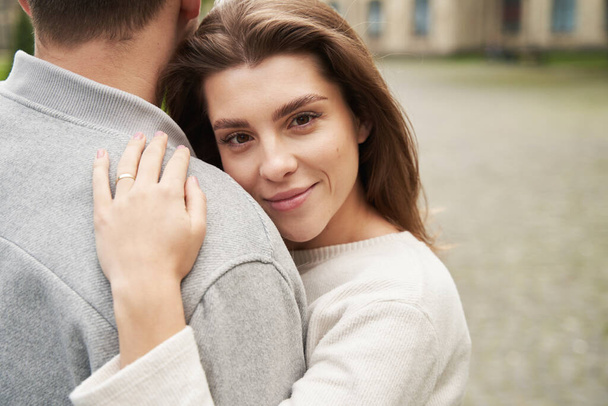 Mujer sonriente enamorada apoyada en el hombro del novio y mirando a la cámara mientras tiene una cita romántica al aire libre - Foto, Imagen