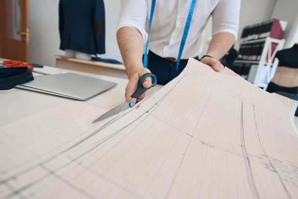 Low angle close up cropped head of male dressmaker cutting paper sketches of new designes at desk with notebook - 写真・画像