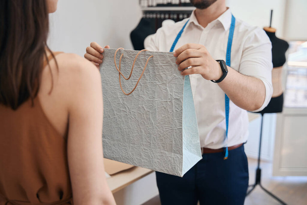 Cropped head of male dressmaker giving paper bag with ready order for female customer in tailor office - Foto, afbeelding