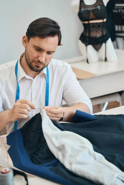 Mannelijke naaister zit aan tafel en gebruikt naald en draad voor het naaien van handgemaakte jas - Foto, afbeelding