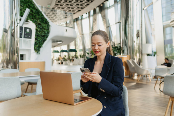 A female realtor in a blazer is typing on a keyboard on the cell phone. A woman is sitting at the table in the modern lobby. A female IT developer near a laptop in a contemporary office interior. - Foto, immagini