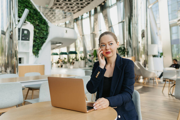 A female realtor in a blazer is having a conversation on the cell phone. A woman is sitting at the table in the modern lobby. A female IT developer near a laptop in a contemporary office interior. - Zdjęcie, obraz