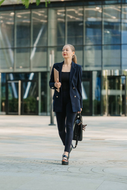 A full length photo of a female realtor in a blazer who is strolling with a laptop between skyscrapers. A businesswoman is leaving her office in a downtown. - 写真・画像