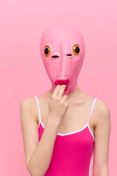 Μια αστεία γυναίκα με ροζ μάσκα ψαριού στο κεφάλι έβαλε τα δάχτυλά της στο στόμα της με ροζ ρούχα σε ροζ φόντο. Η έννοια της σύγχρονης φωτογραφίας τέχνης. Υψηλής ποιότητας φωτογραφία - Φωτογραφία, εικόνα