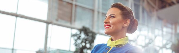 Fröhliche Stewardess in Stewardess-Uniform schaut weg und lächelt, während sie die Arme verschränkt hält - Foto, Bild
