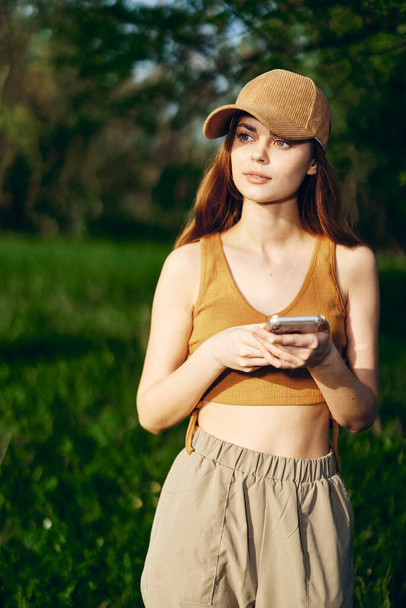A young woman stands with her phone in her hands and smiles in the nature in the sunlight. Freelancer Lifestyle. High quality photo - Photo, Image