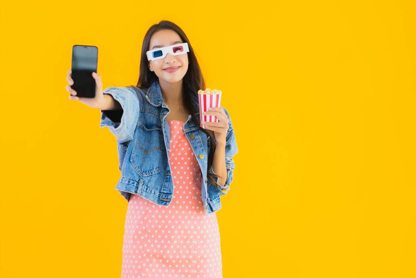 Портрет красивая молодая азиатская женщина наслаждаться счастливым с телефоном попкорн и смотреть фильм на желтом изолированном фоне
 - Фото, изображение