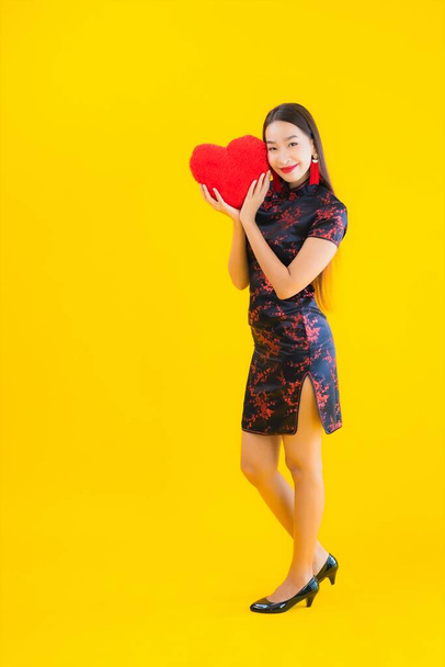 Portrait beautiful young asian woman wear chinese dress show heart shape pillow on yellow isolated background - Foto, afbeelding