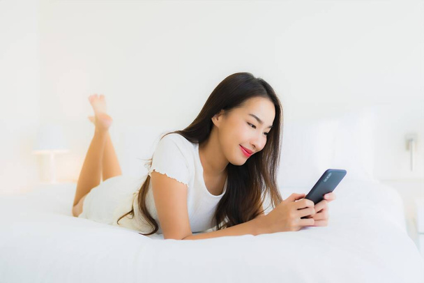 Портрет красива молода азіатка використовує смартфон на ліжку з білою подушкою в інтер'єрі спальні
 - Фото, зображення