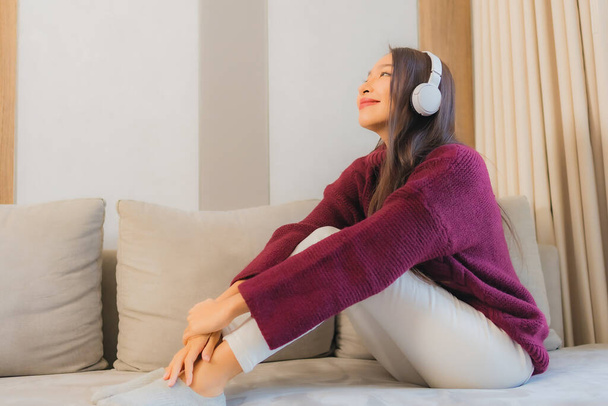Portré gyönyörű fiatal ázsiai nő használja fejhallgató hallgatni zenét kanapén a nappaliban belső - Fotó, kép
