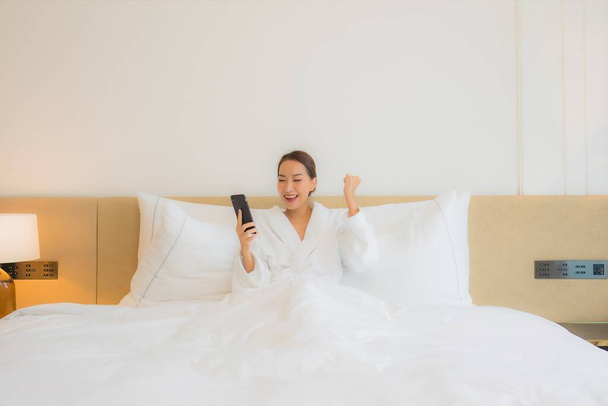 Retrato hermosa joven mujer asiática uso inteligente teléfono móvil en la cama en el interior del dormitorio - Foto, Imagen
