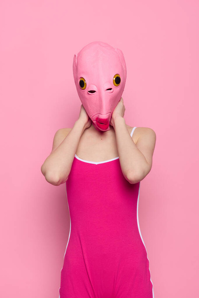 Γυναίκα με στολή ψαριού για τις Απόκριες ποζάρει σε ροζ φόντο με μια τρελή τρομακτική στολή με μια ροζ μάσκα σιλικόνης στο κεφάλι της. Υψηλής ποιότητας φωτογραφία - Φωτογραφία, εικόνα