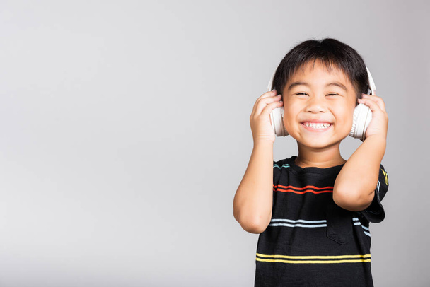 Little cute kid boy 5-6 years old listen music wear wireless headphones in studio shot isolated on white background, happy Asian children holding headset smiling listening audio, entertainment concept - Фото, изображение