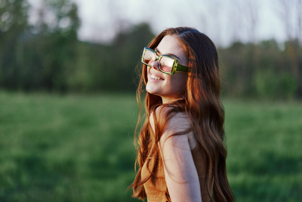 Жінка в сонцезахисних окулярах в парку посміхається і дивиться на захід сонця, крупним планом портрет, копіює простір на задньому плані. Високоякісна фотографія - Фото, зображення