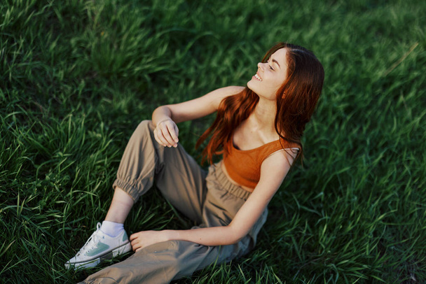 Top näkymä nuori punapäinen nainen istuu luonnossa vihreällä ruoholla ja hymyilee ja peittää kasvonsa hiuksillaan hämmennystä. Laadukas kuva - Valokuva, kuva