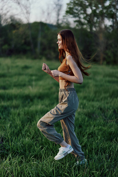 Μια γυναίκα με μακριά κόκκινα μαλλιά γυμνάζεται και τρέχει στο πράσινο γρασίδι στο πάρκο με φόρμες και αθλητικά παπούτσια. Υψηλής ποιότητας φωτογραφία - Φωτογραφία, εικόνα