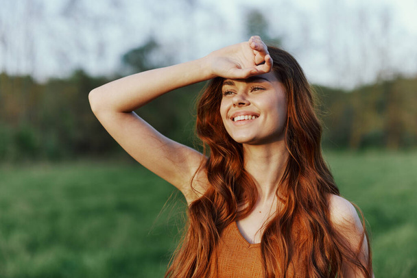 Молода руда жінка посміхається на заході сонця в зеленому парку і тримає руку на волоссі. Високоякісна фотографія - Фото, зображення