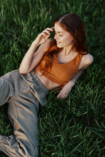 Молода жінка з довгим рудим волоссям посміхається на зеленій траві в парку, розслабляючись після спортивних тренувань під час заходу сонця. Високоякісна фотографія - Фото, зображення