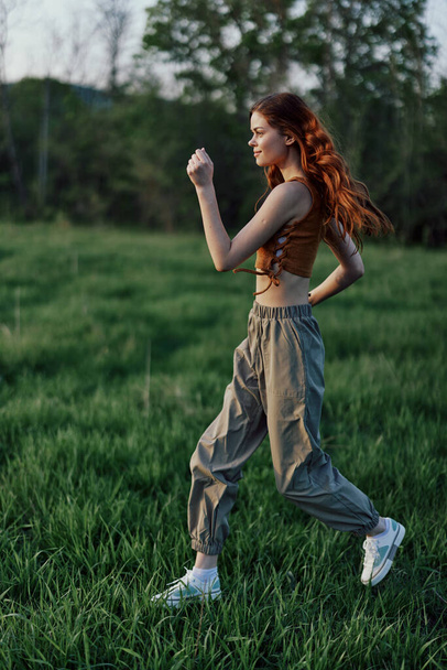 Una mujer con el pelo rojo largo se ejercita y corre sobre la hierba verde en el parque en pantalones de chándal y zapatillas de deporte. Foto de alta calidad - Foto, imagen