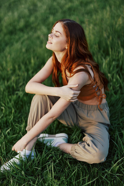 Güzel kızıl saçlı bir kadın bahçedeki yeşil çimenlerde oturur ve güneşin batışına bakarak çiçek açan doğayı seyreder. Yüksek kalite fotoğraf - Fotoğraf, Görsel