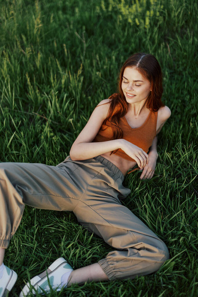 A woman lying on the green grass in green pants and an orange top, smiling in the sunset summer sun in nature. High quality photo - Photo, Image