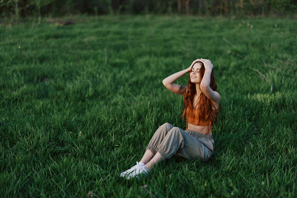 Na trávě v parku sedí zrzavá žena s dlouhými, větrem rozcuchanými vlasy a usmívá se, tvář jí ozařuje světlo zapadajícího slunce. Koncept harmonie s přírodou venku. Kvalitní fotografie - Fotografie, Obrázek