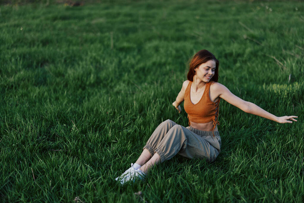 Nuori punapäinen nainen istuu puistossa vihreällä ruoholla kädet ojennettuina eri suuntiin ja hymyillen laskevan auringon valossa. Laadukas kuva - Valokuva, kuva