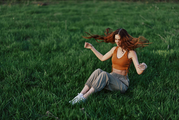 Rudowłosa kobieta z długimi, wietrznymi włosami siedzi na zewnątrz na trawie w parku i uśmiecha się, a zachód słońca oświetla jej twarz. Koncepcja harmonii z naturą na zewnątrz. Wysokiej jakości zdjęcie - Zdjęcie, obraz
