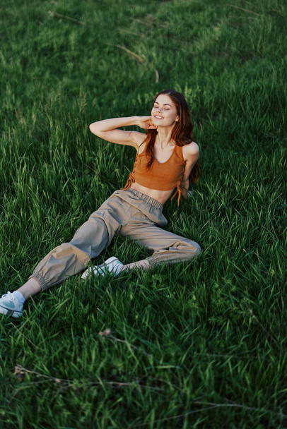 Nainen makaa tuoreella kevään vihreällä ruoholla puistossa ja nauttii rentouttavasta auringonlaskun katselusta. Laadukas kuva - Valokuva, kuva