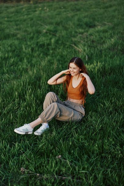 Μια γυναίκα που απολαμβάνει την ύπαιθρο κάθεται στο πάρκο στο πράσινο γρασίδι με casual ρούχα με μακριά μαλλιά που ρέει, φωτίζεται από τον λαμπερό καλοκαιρινό ήλιο χωρίς κουνούπια. Υψηλής ποιότητας φωτογραφία - Φωτογραφία, εικόνα
