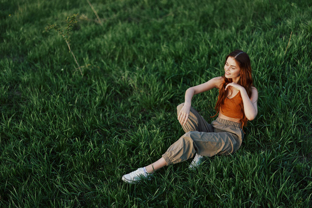 Uma mulher bonita com cabelo longo e encaracolado vermelho senta-se no parque no verão na grama verde na luz do pôr do sol. Foto de alta qualidade - Foto, Imagem