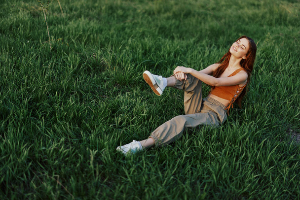 Руда жінка сидить у парку на зеленій траві в помаранчевій верхній частині, зелених штанах та кросівках і дивиться на місце літнього сонця. Високоякісна фотографія - Фото, зображення
