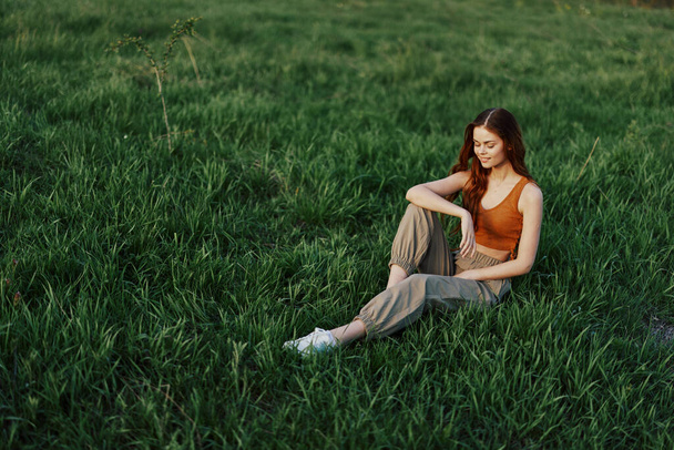 La pelirroja se sienta en el parque en la hierba verde vistiendo una camiseta naranja, pantalones verdes y zapatillas de deporte y mira hacia el atardecer del verano. Foto de alta calidad - Foto, Imagen