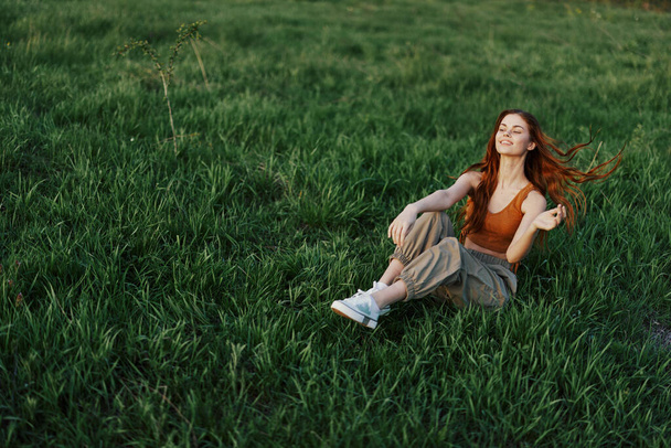 A mulher ruiva se senta no parque na grama verde usando um top laranja, calças verdes e tênis e olha para o pôr do sol de verão. Foto de alta qualidade - Foto, Imagem