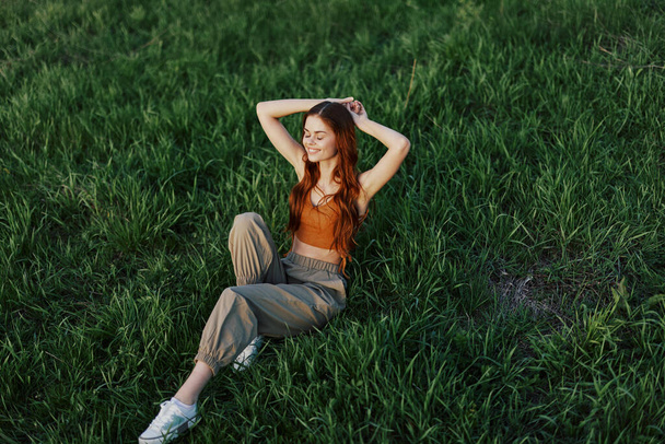 Una hermosa mujer con el pelo largo y rizado se sienta en el parque en el verano en la hierba verde en la luz del atardecer. Foto de alta calidad - Foto, Imagen