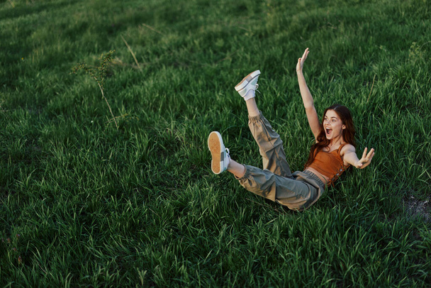 Μια νεαρή γυναίκα παίζει στο πάρκο μαζί μας στο γρασίδι και πέφτει στο έδαφος χαμογελώντας χαρούμενα στο φως του ήλιου. Ξεκούραση στη φύση, αρμονία με το σώμα. Υψηλής ποιότητας φωτογραφία - Φωτογραφία, εικόνα