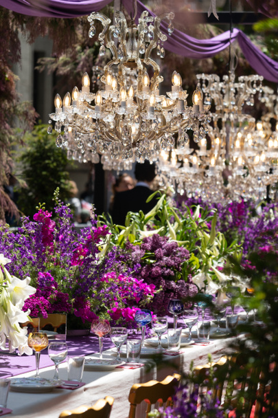 ガラスのゴブレットやメガネ、ホワイトプレート、豪華な結婚式や誕生日パーティーのためのお祝いのテーブルセッティングのためのカトラリー。新鮮な紫色のピンク色の花や花束と装飾。ソフトフォーカス. - 写真・画像
