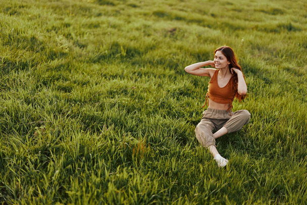 Солнечный свет летнего вечера освещает красивую молодую рыжую женщину, сидящую на траве в зеленых штанах и оранжевом топе и улыбающуюся солнцу. Высокое качество фото - Фото, изображение