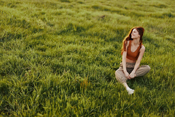 Young woman with red hair is happy and smiling sitting on the fresh grass in the park in the sun. High quality photo - Photo, Image