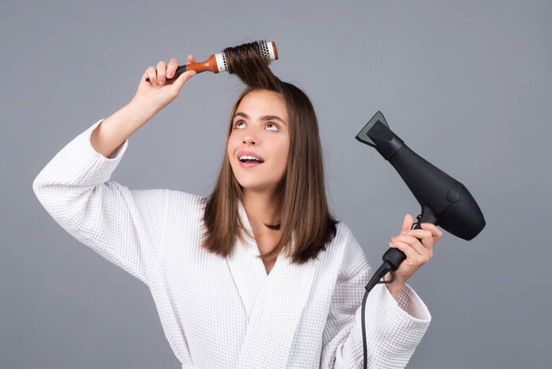 Woman in bathrobe combing hair, dryer hair with hairdryer. Portrait of female model with a comb brushing hair. Girl with hairbrush, hair care and beauty. Morning routine - Foto, Imagem