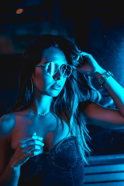 Portrait of a brunette girl at night in the city with blue leds and blue glasses, looking at the camera - Фото, изображение
