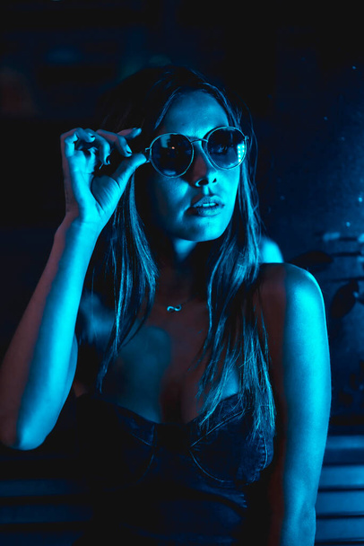 Portrait of a brunette girl at night in the city with blue leds and blue glasses, looking at the camera - Fotó, kép